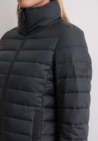 Svart dunjakke med høy krage, horisontalt quiltet design, glidelås lukking og strukturert stoff med en logo-patch på ermet.