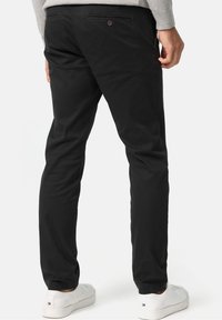 Black cotton trousers with a straight fit, featuring back pockets with button closures. Worn with white sneakers and a gray top.