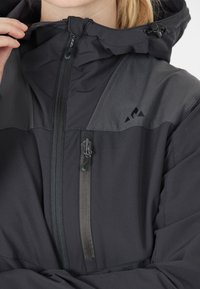 Svart utomhusjacka med en slät textur, framzipper och justerbar huva med en kontrasterande grå panel och logotypdetalj.