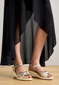 Metallic gold espadrille sandals with knotted straps, featuring a wedge heel and ankle strap. Background is a flowing black sheer fabric.