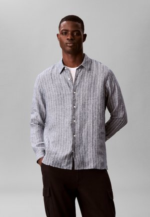 Mann trägt ein grau-weiß gestreiftes Hemd über einem weißen T-Shirt, mit schwarzer Hose, steht mit teilweise in den Taschen steckenden Händen.