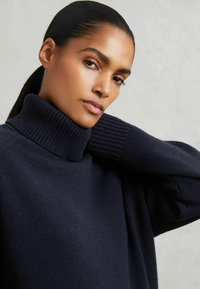 Pull en maille marine avec un col roulé chunky et des poignets larges côtelés, présentant une texture douce et une silhouette décontractée.