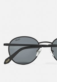 Gafas de sol de metal negro con lentes circulares, tono gris y almohadillas nasales transparentes. Presentan un logotipo dorado en la varilla. Diseño de marco delgado.