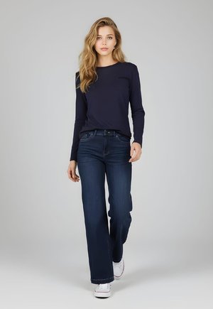 Mujer joven caminando hacia la cámara, vestida con una camisa azul marino de manga larga, jeans azul oscuro y zapatillas blancas, sobre un fondo gris.