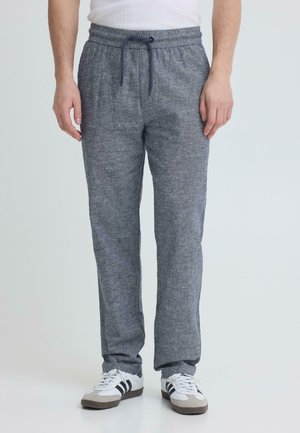 Mand iført grå sweatpants med snørre og hvide sneakers med sorte striber, stående mod en neutral baggrund.