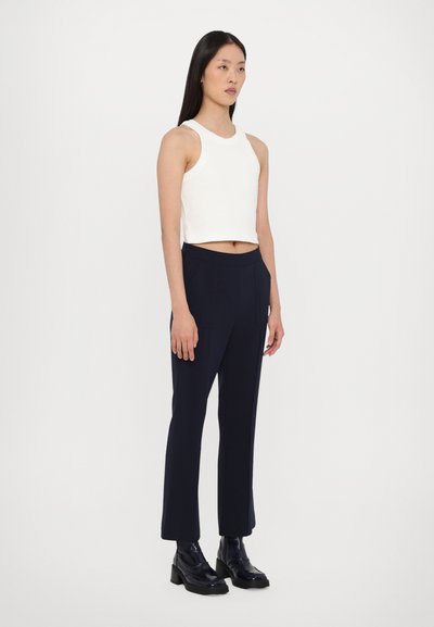 Hvid, ærmeløs crop top med en glat tekstur; parret med navy høj talje bukser og sorte glansfulde ankelstøvler. Simpel design.