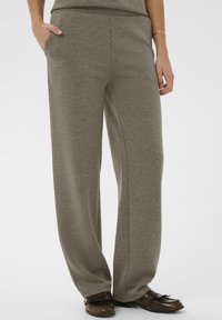 Osoba oblečená v voľných taupe nohaviciach s bočnými vreckami a hnedými koženými mokasínami, stojaca na jednoliatom pozadí.