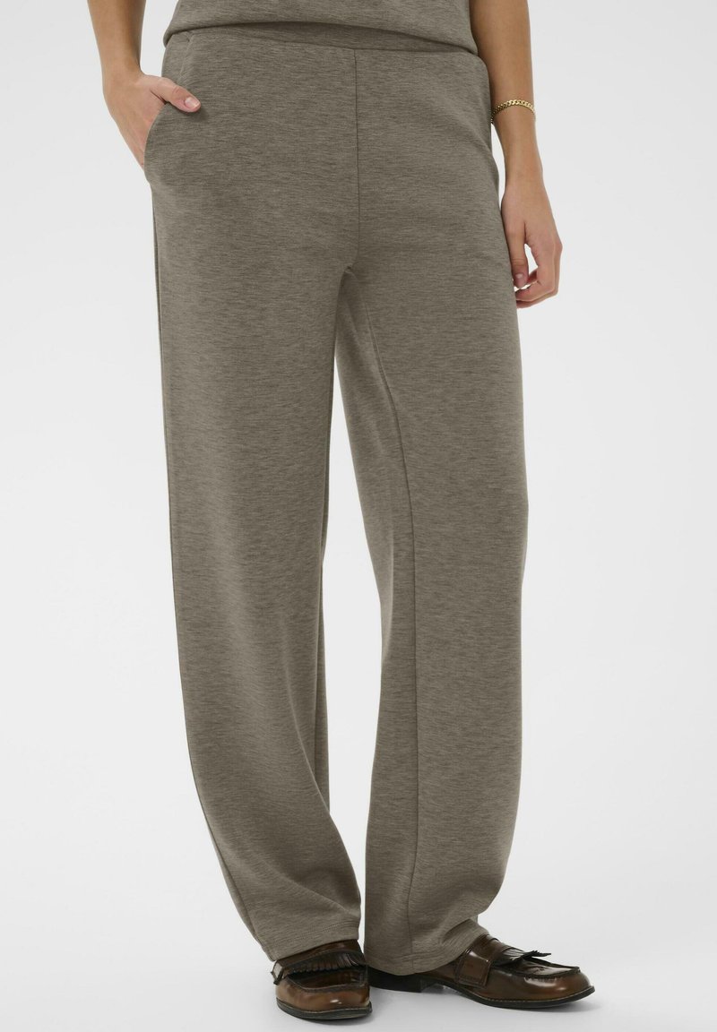 Osoba oblečená v voľných taupe nohaviciach s bočnými vreckami a hnedými koženými mokasínami, stojaca na jednoliatom pozadí.