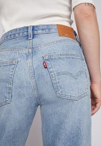 Personne portant un jean Levi's bleu clair et un haut blanc côtelé, montrant les poches arrière et le patch en cuir de la marque sur la ceinture.