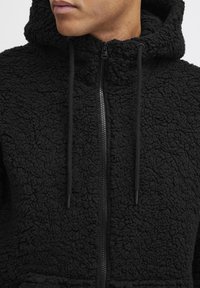 Black fleece zip-up hoodie featuring a textured, fur-like exterior, adjustable drawstrings, and a front zipper.