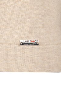 Maglione beige lavorato a maglia con una texture morbida, dotato di un'etichetta logo metallica che riporta la scritta "FELIX HARDY" con accenti in nero, rosso e blu.