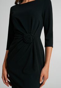 Robe noire en tissu lisse avec des manches trois-quarts, une silhouette cintrée et un design torsadé à la taille agrémenté d'un détail en forme de rose.