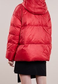 Rød dunjakke med hætte, med en skinnende overflade, vandrette syninger og en løs pasform. Båret med en sort nederdel.