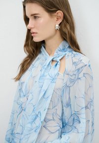 Lyseblå gennemsigtig bluse med blomsterdesign, der har en knyttet halsbånd og åbne skulderudskæringer. Glat tekstur med fine detaljer.