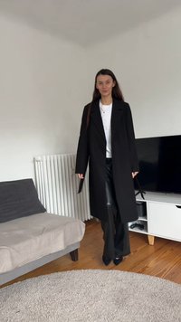 Casaco longo preto sobre uma camisa branca, combinado com calças de couro pretas e saltos altos. Em pé contra uma parede clara e mobiliário moderno.