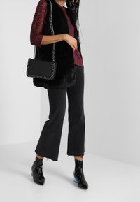 Schwarze Kunstfellweste über einem burgunderfarbenen Oberteil, kombiniert mit schwarzen, taillierten Jeans und glänzenden schwarzen Ankle-Boots mit blauen Absatzakzenten. Silberne Kettenhandtasche.