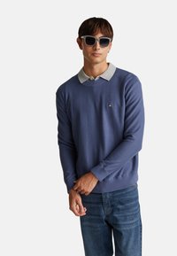 Niebieski sweter dzianinowy z szarym kołnierzem, wyposażony w prążkowane mankiety i dół. Małe haftowane logo na piersi. Noszony z dżinsami i okularami przeciwsłonecznymi.