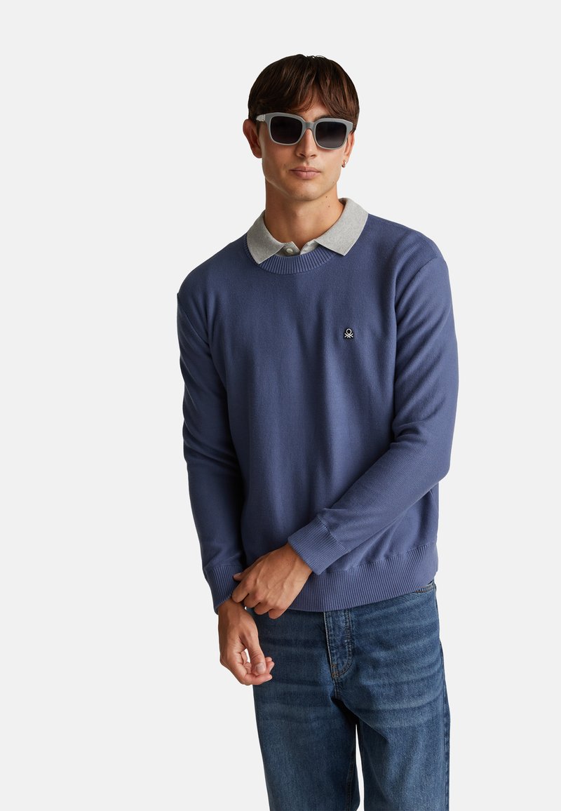Niebieski sweter dzianinowy z szarym kołnierzem, wyposażony w prążkowane mankiety i dół. Małe haftowane logo na piersi. Noszony z dżinsami i okularami przeciwsłonecznymi.