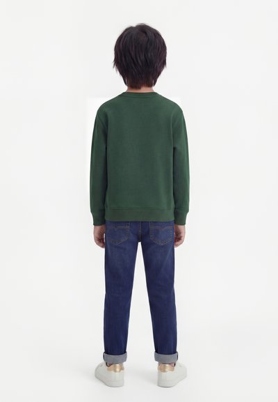 Παιδί που στέκεται με την πλάτη στραμμένη, φοράει σκουρό πράσινο φούτερ, μπλε τζιν με γυρισμένα μπατζάκια και λευκά αθλητικά με χρυσές λεπτομέρειες.