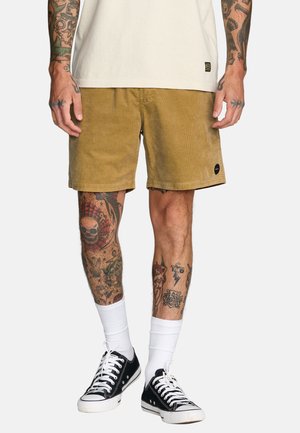 Tan ribfluwelen shorts met een relaxte pasvorm, voorzien van een rechte zoom en een klein logoplaatje. Gecombineerd met zwarte sneakers en witte sneakersokken.