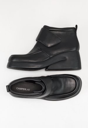 Bottines en cuir noir avec talons compensés et larges bandes velcro, une botte vue de côté, l’autre vue de dessus avec semelle intérieure « CAMPERLAB ».
