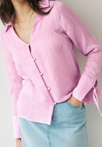 Chemise boutonnée rose clair avec texture à côtes verticales, manches longues et décolleté en V. Dotée d'accents de boutons et de liens sur les poignets.