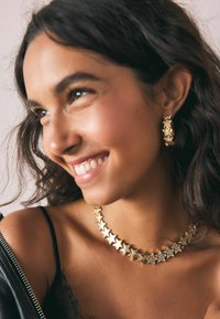 Kuldne tähekujuline kaelakee vahelduvate läikivate ja rhinestone'idega kaunistatud tähtedega, kantuna musta topi ja nahktagi all.