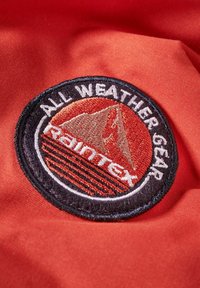 Okrągła naszywka haftowana na czerwonym tle, z napisem "ALL WEATHER GEAR", motywem górskim oraz brandingiem "Raintex" w ciemnym obrysie.