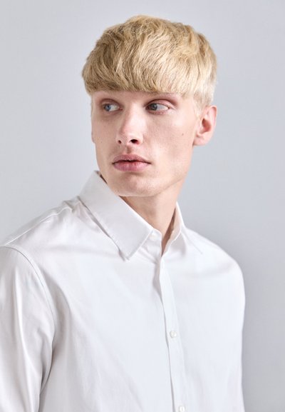 Chemise blanche à boutons en tissu de coton lisse, avec un col classique et une coupe ajustée, portée par un mannequin aux cheveux blonds courts.