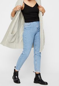 Woman wearing a cream knit cardigan, black sleeveless top, light blue jeans, and black ankle boots with a small heel.