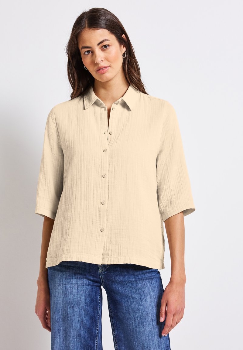 Camicia button-up beige chiaro in tessuto testurizzato, maniche a 3/4 e colletto, abbinata a jeans blu. Dettagli di cucitura sottili presenti.