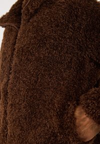 Close-up van een zacht, textuur rijk bruin fleece jacket met zichtbare rits en een hand in de zijzak gestoken.