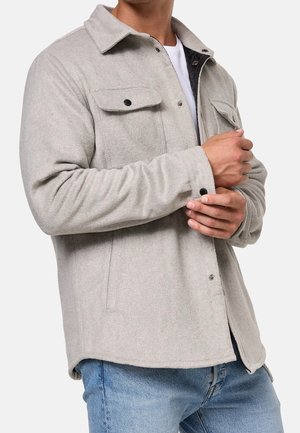 Homme ajustant le poignet d'une veste boutonnée gris clair portée par-dessus une chemise blanche et un jean bleu sur fond uni.