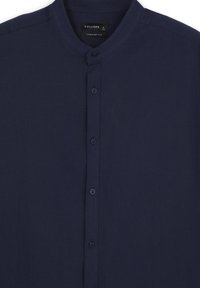 Chemise bleu marine à boutons avec col montant, dotée d'une texture lisse et d'une patte de boutonnage cachée. L'étiquette indique "CALLIOPE", taille L.