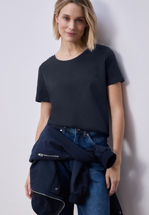 Mujer rubia con camiseta negra de manga corta y vaqueros azules, con chaqueta azul marino atada a la cintura, de pie contra una pared clara.