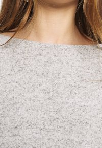 Světlý šedý pletený top s texturovaným vzorem, výstřihem v loděním stylu a měkkou látkou, částečně viditelný od oblasti hrudi.