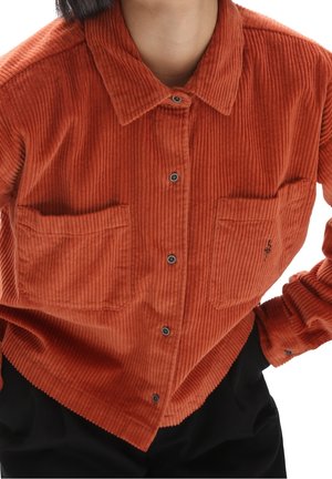 Camisa de pana color óxido con botones y dos bolsillos en el pecho, usada sobre pantalones negros, mostrada desde el cuello hasta la cintura.