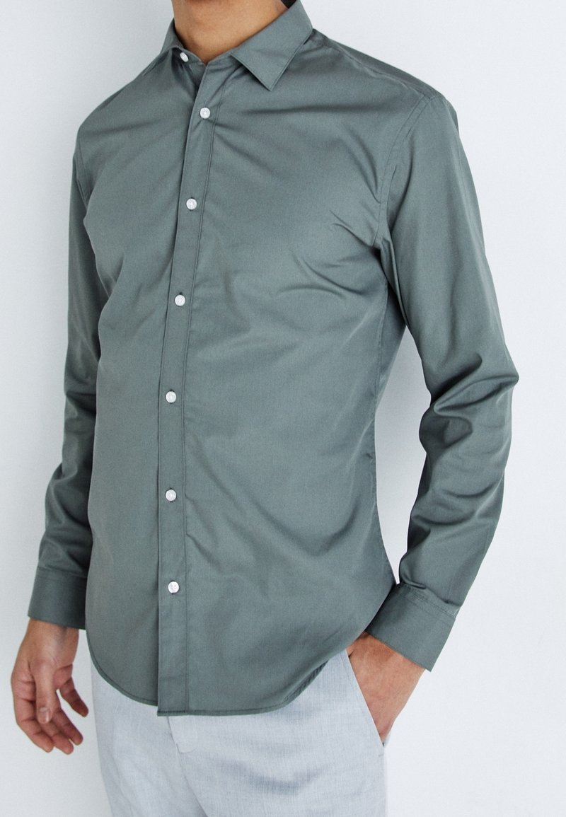 Camicia verde a maniche lunghe in un tessuto liscio, con colletto classico e bottoni bianchi, abbinata a pantaloni grigio chiaro.