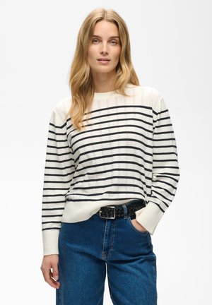 Femme aux longs cheveux blonds porte un pull blanc à rayures noires horizontales et un jean bleu, debout avec une main dans la poche.
