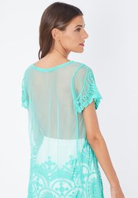 Cubierta turquesa transparente con mangas acampanadas y un intrincado patrón bordado en el dobladillo. Tela ligera con un diseño fluido.