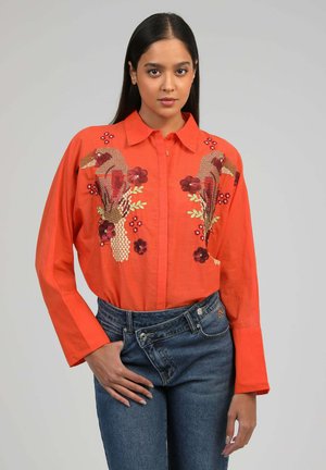 Femme portant une chemise orange avec broderies d'oiseaux et de fleurs, associée à un jean bleu taille haute asymétrique, debout devant un fond uni.
