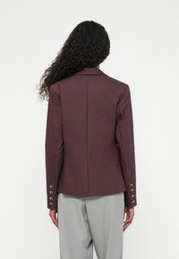 Blazer bordeaux con una vestibilità su misura, rever a coda di rondine e bottoni silver sulle maniche. Indossato con pantaloni grigi chiari a righe. Texture liscia.