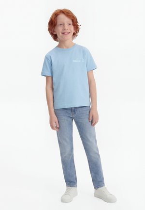 Red-haired boy wearing light blue t-shirt, blue jeans, and white sneakers, standing and smiling, facing slightly left on white background.