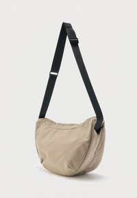 Μπεζ τσάντα χιαστί από μαλακό ύφασμα με φαρδιά, ρυθμιζόμενη μαύρη λουρίδα σε απλό φωτεινό φόντο.