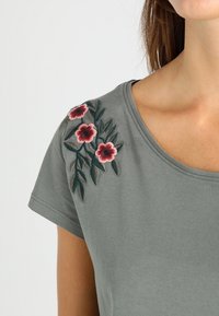 Grå kortærmet T-shirt med pink og mørkegrøn blomsterbroderi på venstre skulder. Blødt bomuldsstof med rund halsudskæring.