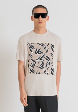 T-shirt beige avec un grand motif carré de feuilles noires sur un fond clair. Le tissu est lisse, associé à un pantalon sombre et des lunettes de soleil surdimensionnées.
