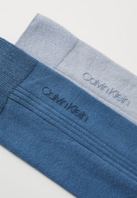 Două perechi de șosete în nuanțe de albastru și gri deschis, cu o textură în relief și logo-ul "Calvin Klein" embosat pe manșete.