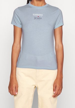 Osoba nosząca jasnoniebieski t-shirt z krótkim rękawem z logo „tommy jeans” i kremowe spodnie sztruksowe, stojąca z rozluźnionymi rękami wzdłuż ciała.