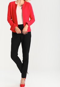 Rode textuurjacket over een lichtgepatteerd top, gecombineerd met zwarte broeken en rode hoge hakken. Minimalistisch ontwerp met een gestroomlijnde silhoutte.