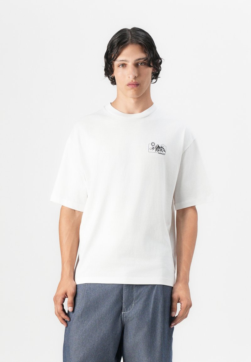 T-shirt en coton blanc à manches courtes et col ras du cou. Présente un petit graphique noir de montagnes et d'un soleil sur la poitrine gauche.